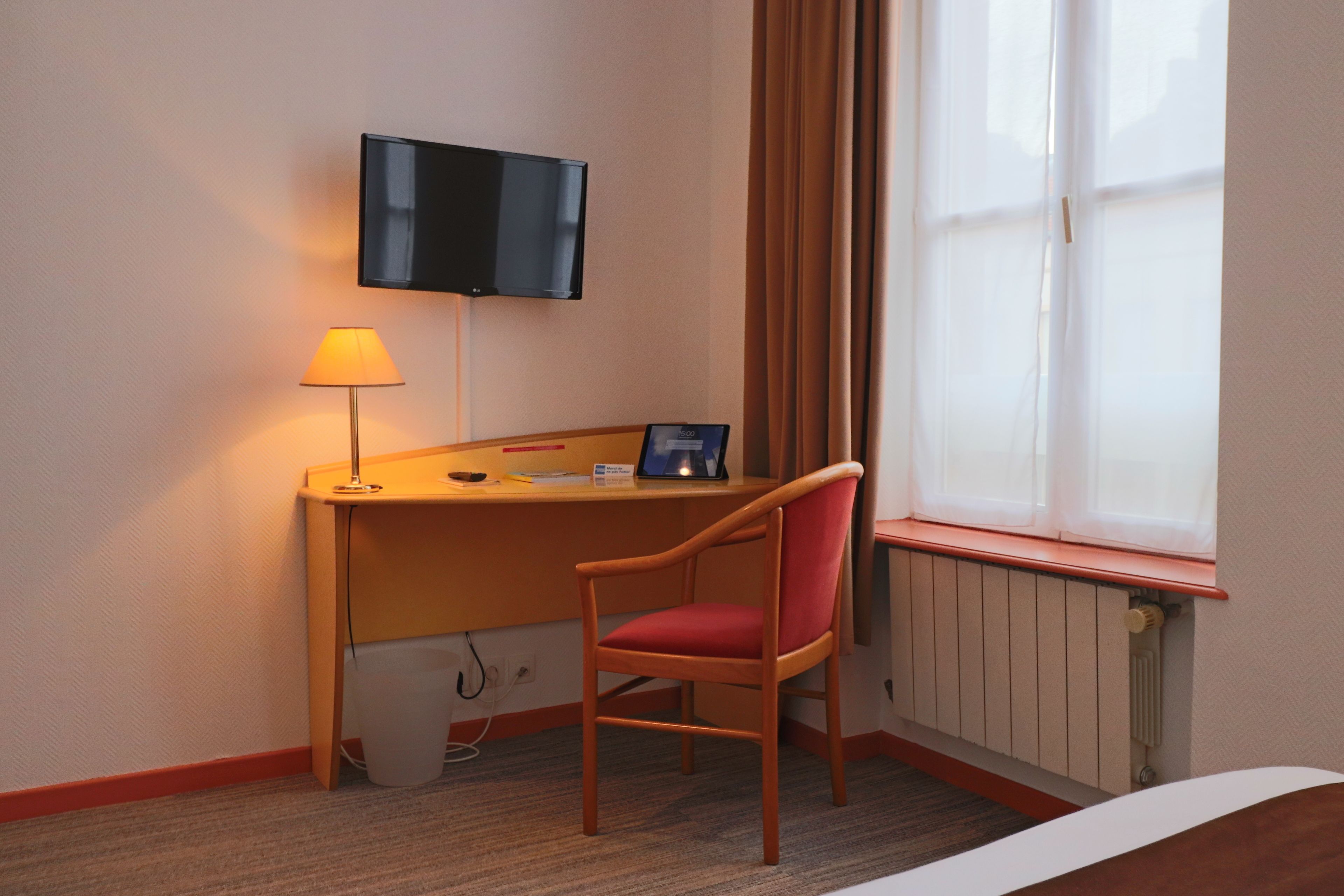 A hotel room with a desk, a chair, a television mounted on the wall, and a window with curtains.