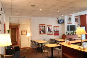 A hotel lobby with multiple tables and chairs, a reception desk, and a television mounted on the wall.