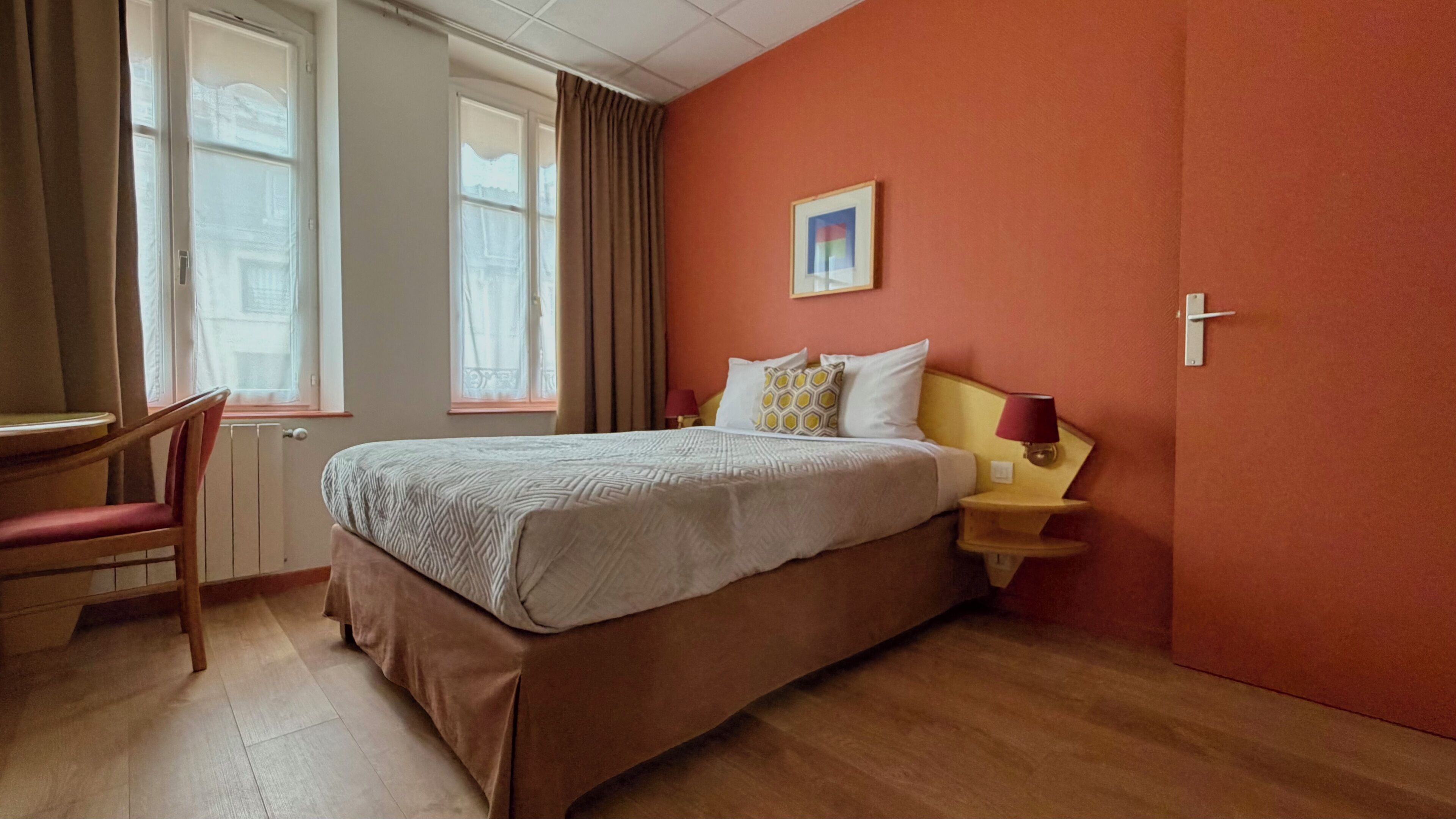 Una habitación de hotel con cama, una silla, una mesita de noche con lámpara y una ventana con cortinas.