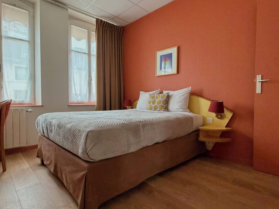 Una habitación de hotel con cama, una silla, una mesita de noche con lámpara y una ventana con cortinas.