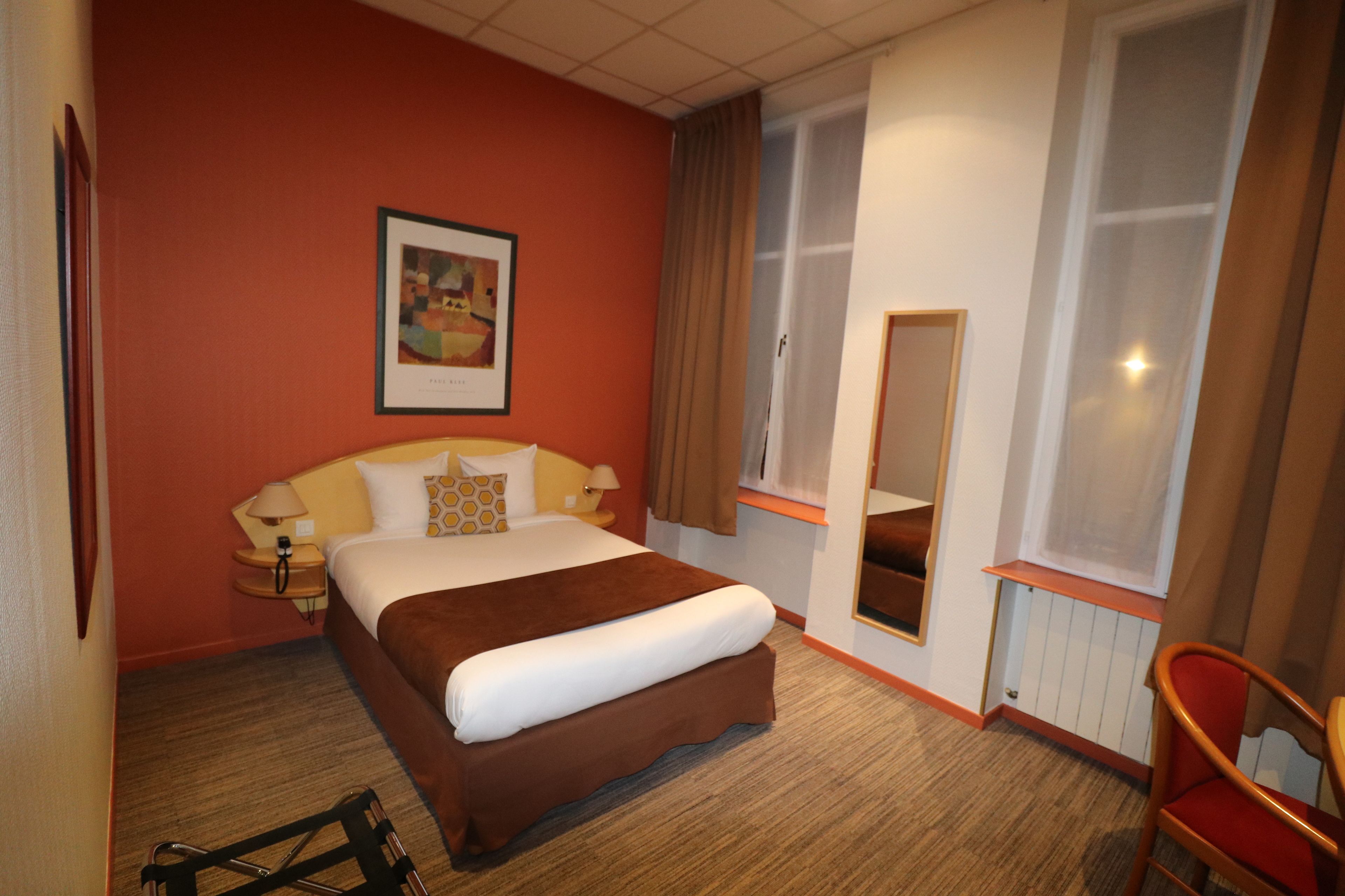 Una habitación de hotel con una cama grande, una mesita de noche con lámpara, una ventana con cortinas e imagen enmarcadas en la pared.