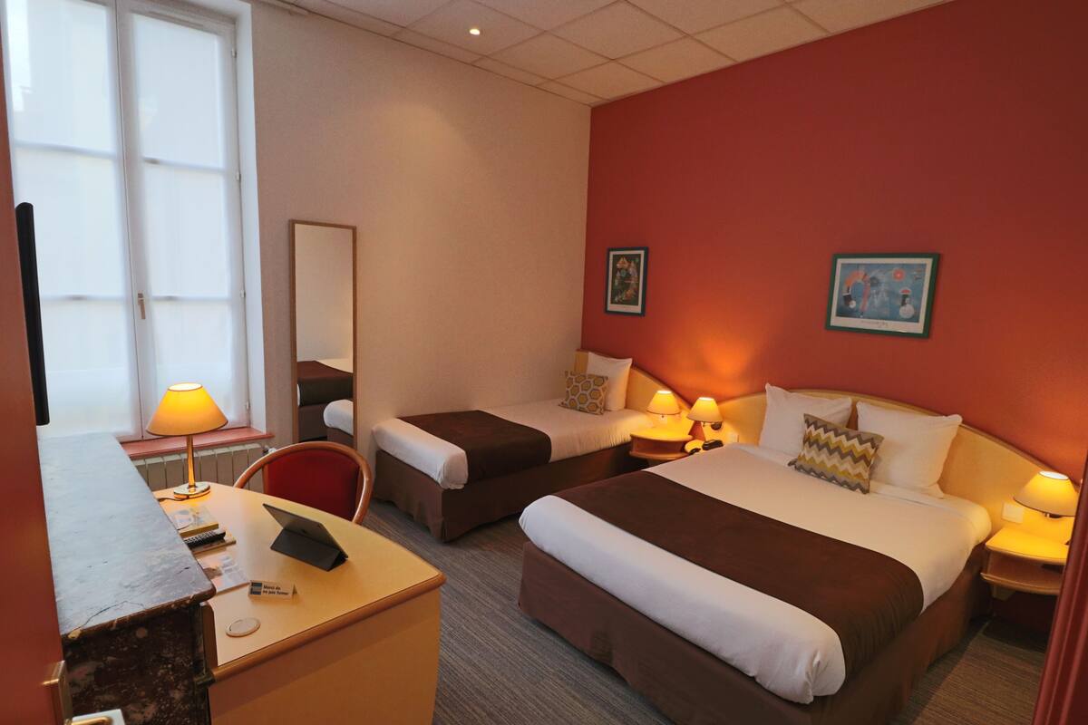 A hotel room with two beds, a desk with a laptop, a chair, and a window with a lamp.