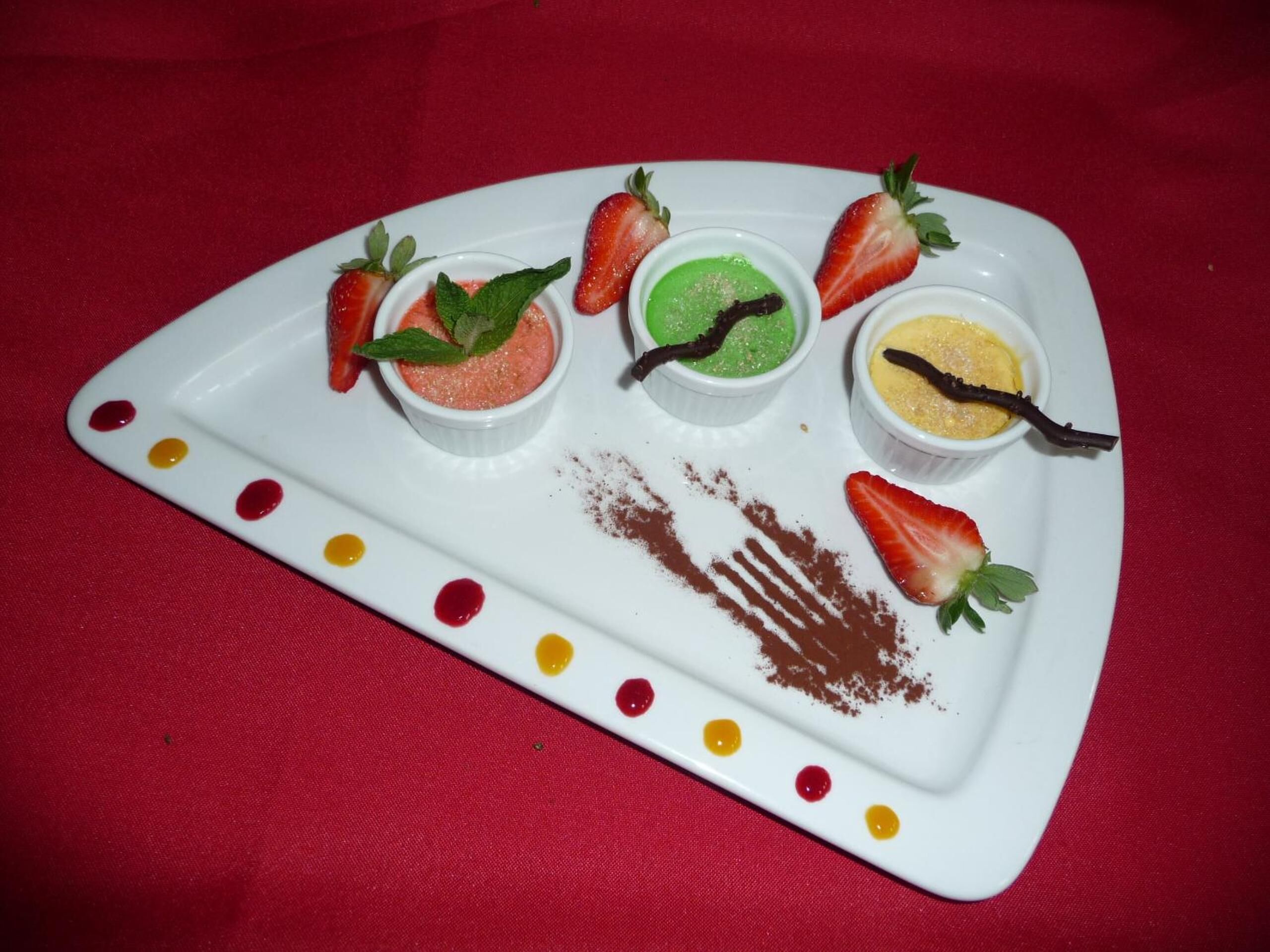Un plato blanco con cuatro vasitos de postre, cada uno decorado con una fresa y una hoja de menta, sobre un mantel rojo.