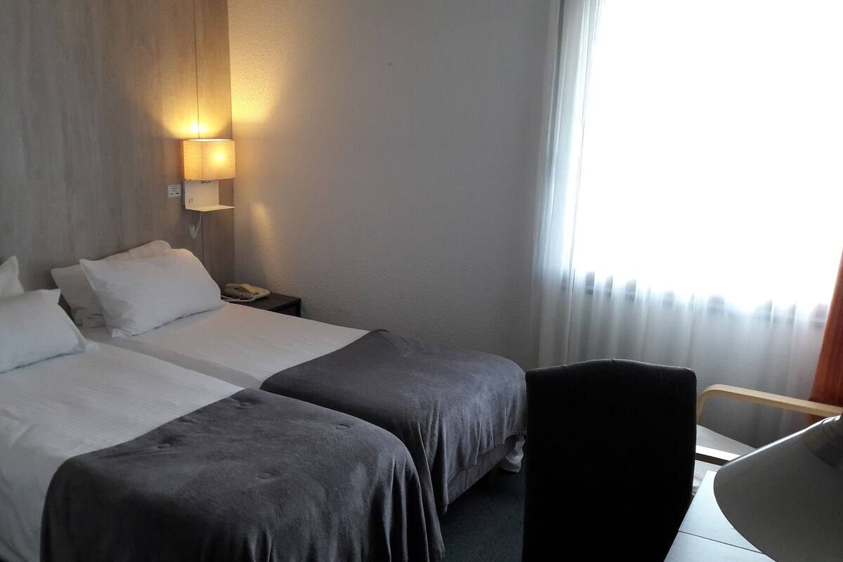 Habitación de hotel con dos camas, una mesita de noche con lámpara, una silla y un ventanal con cortinas.