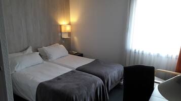 A hotel room with two beds, a bedside table with a lamp, a chair, and a window with curtains.