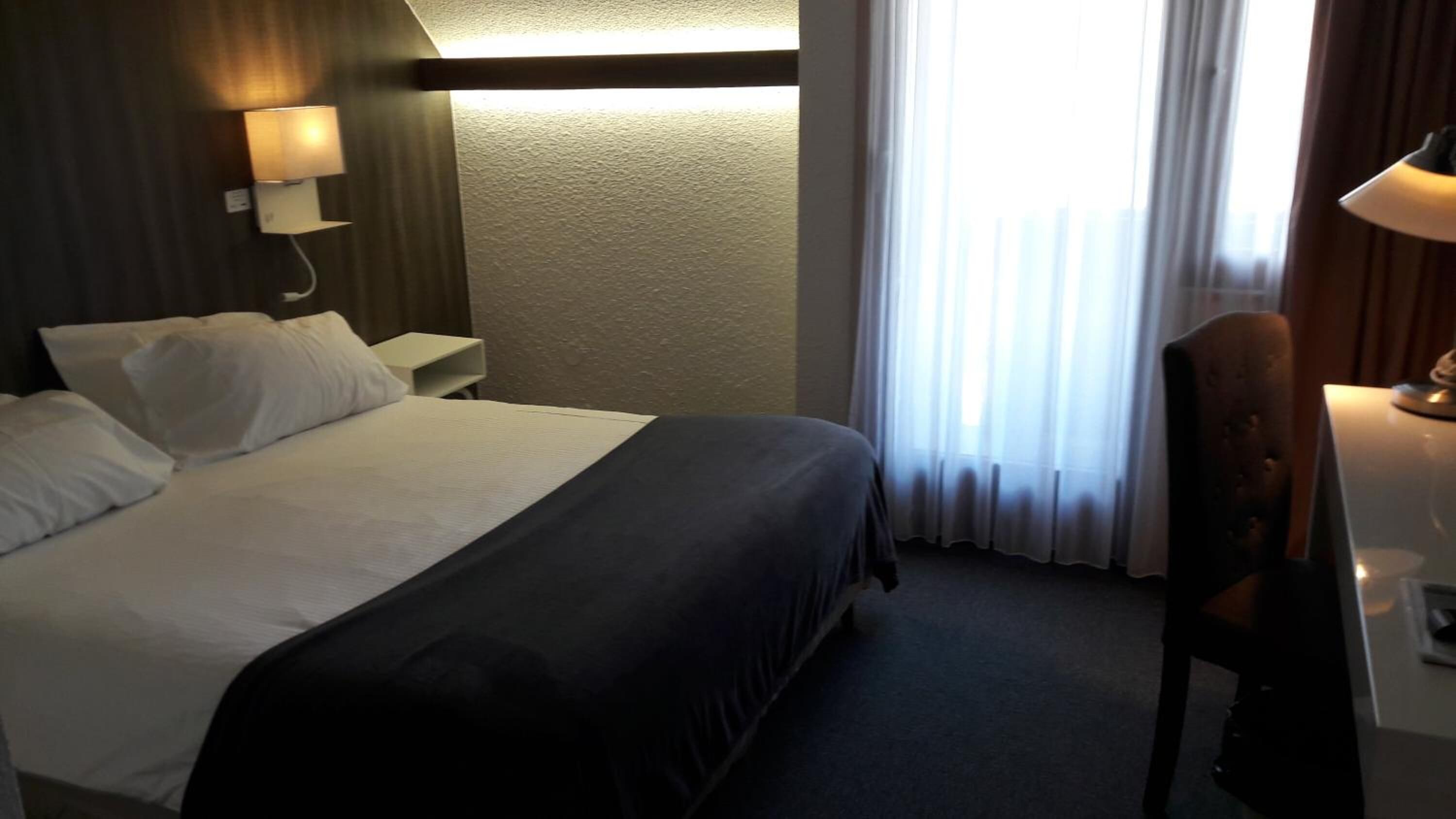 A hotel room with a bed, a nightstand, a lamp, a chair, and a window with curtains.
