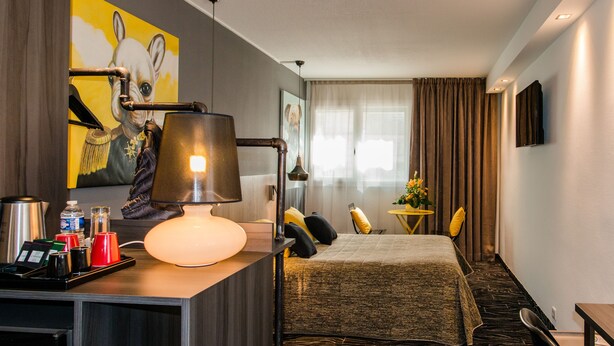 A hotel room with a bed, a desk with a lamp, a coffee maker, and a small table with a vase of flowers.