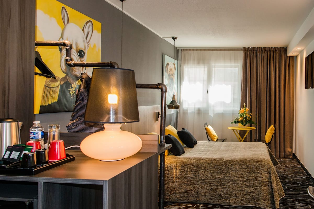 A hotel room with a bed, a desk with a lamp, a coffee maker, and a small table with a vase of flowers.