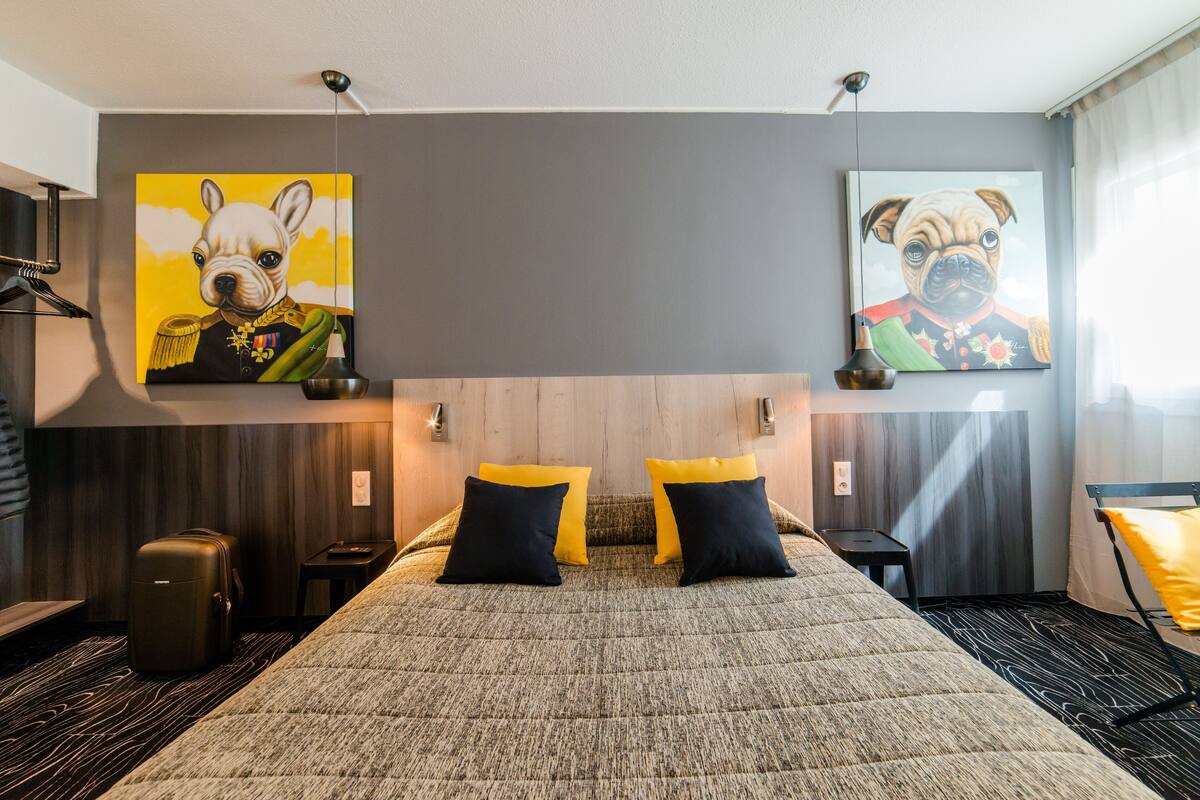 A hotel room with a large bed, two paintings of dogs, a desk, and a chair.