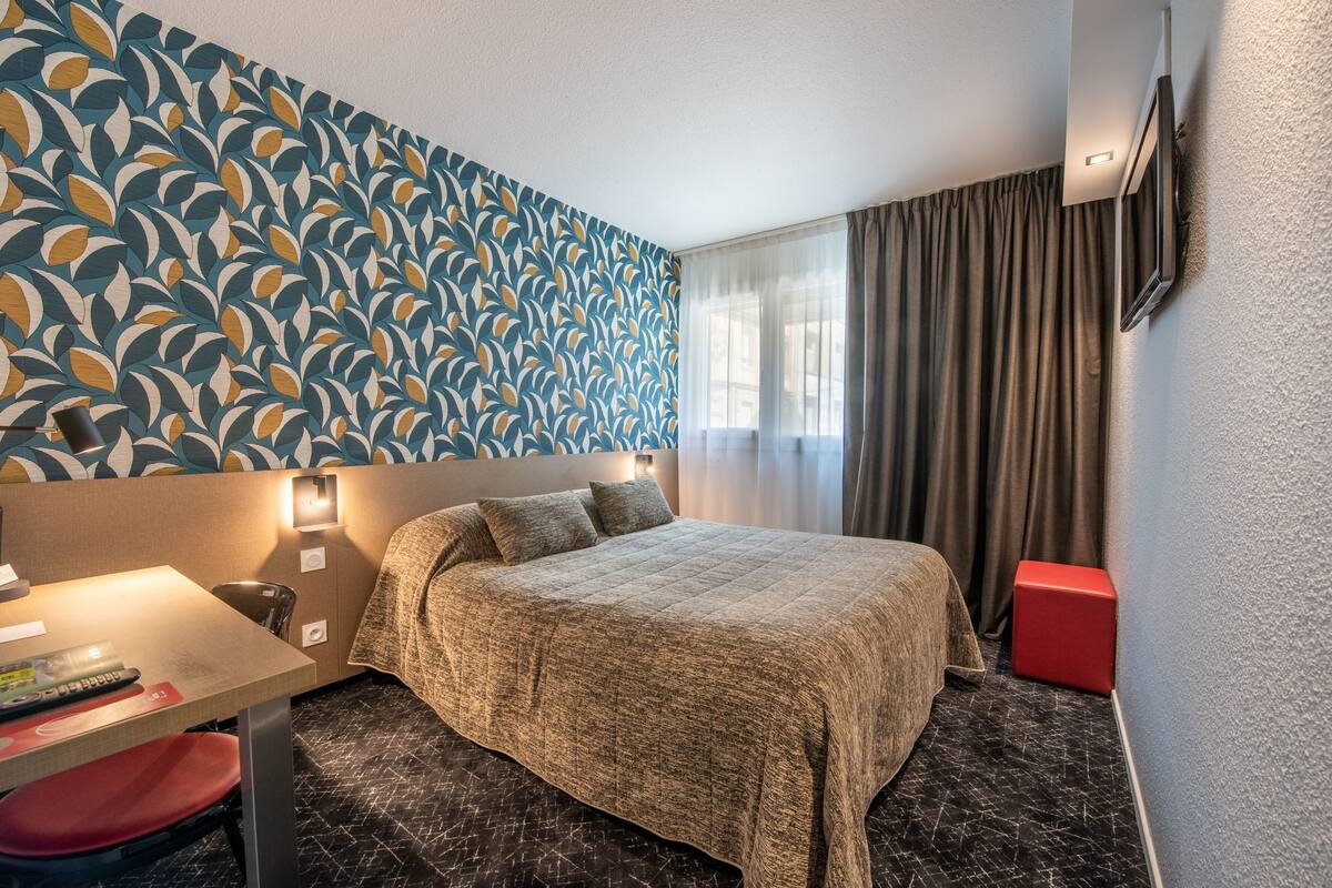 A hotel room with a bed, a desk with a chair, a television mounted on the wall, and a patterned wallpaper.