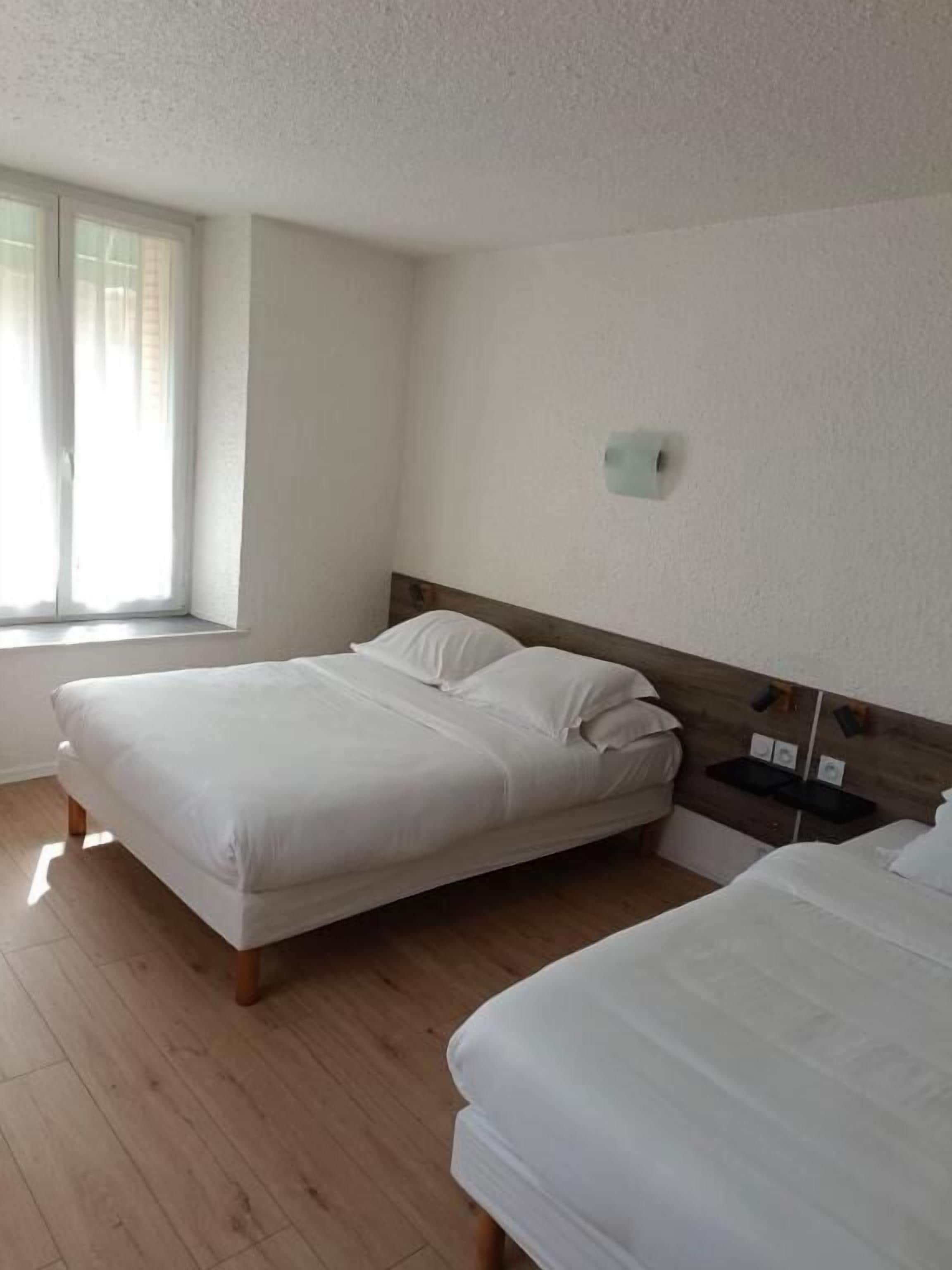 A room with two beds, a window with blinds, a wall-mounted light, and a wooden floor.
