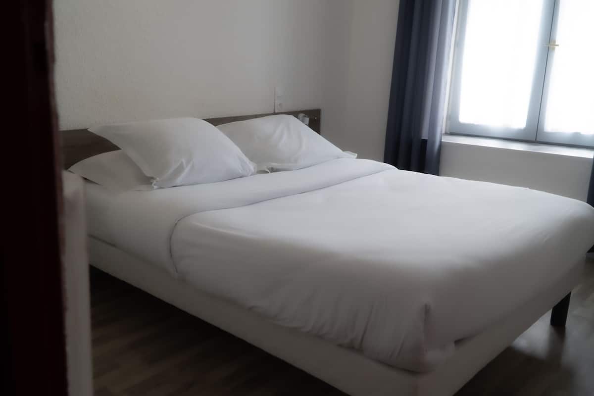 A neatly made bed with white linens in a room with wooden flooring and a window with curtains.