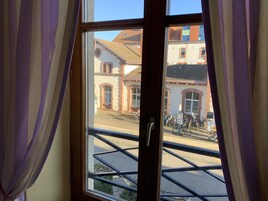 Una ventana con cortinas moradas, vista de un edificio con ventanas de arco y tejado de tejas, y una bicicleta estacionada afuera.