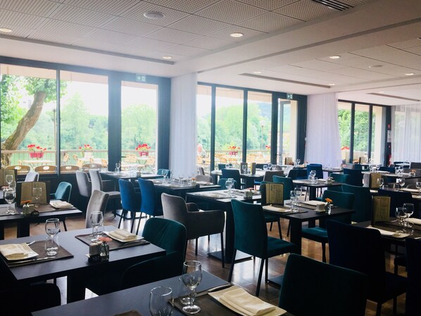 A restaurant with multiple tables and chairs, large windows, and a view of greenery outside.