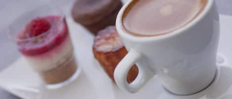 Eine weiße Kaffeetasse auf einem Untertasse mit einem Löffel daneben, und zwei Desserts auf einem weißen Teller.