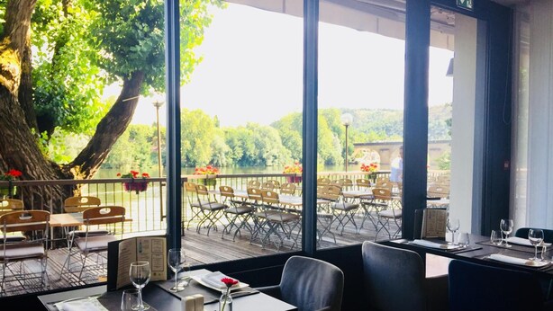 Restaurante con terraza al aire libre, vistas a un lago, un árbol grande y vista del agua.