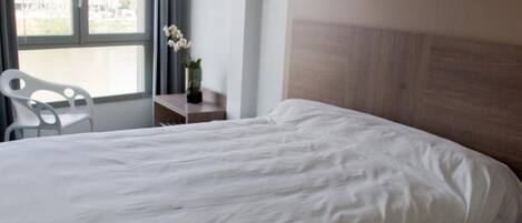 A hotel room with a large bed, a wooden headboard, a small table, a chair, and a window with curtains.