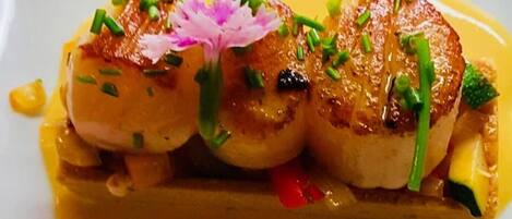 Une assiette de coquilles Saint-Jacques poêlées, garnie de ciboulette et d’une fleur, servie avec une sauce jaune.