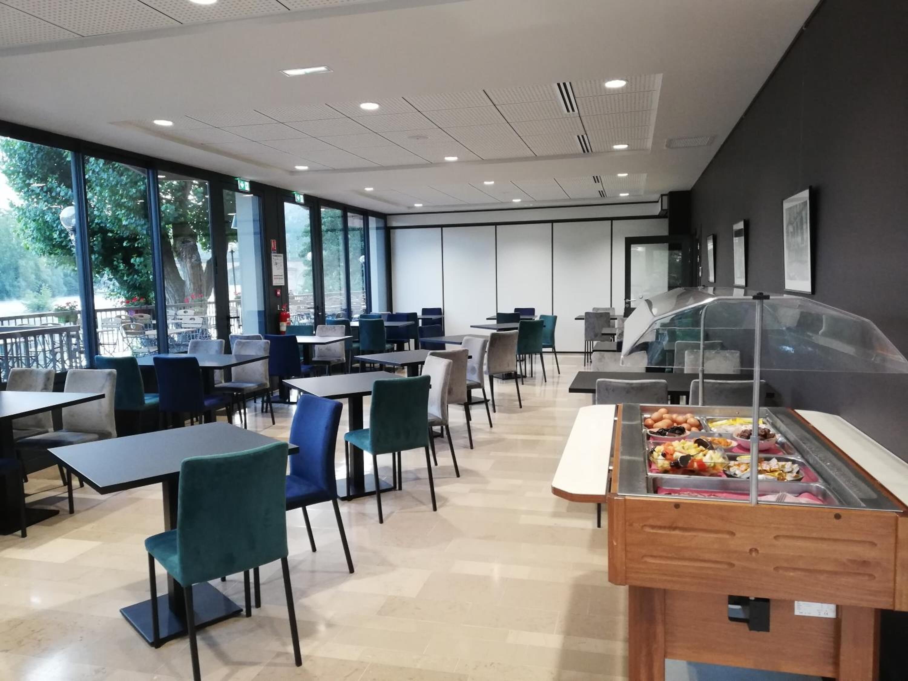 Una cafetería moderna con mesas y sillas, ventanas amplias y una zona de exhibición de alimentos.