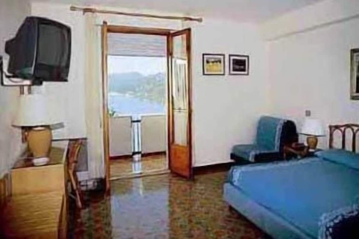Habitación de hotel con cama, televisión, escritorio con lámpara y vista al mar a través de una puerta abierta.