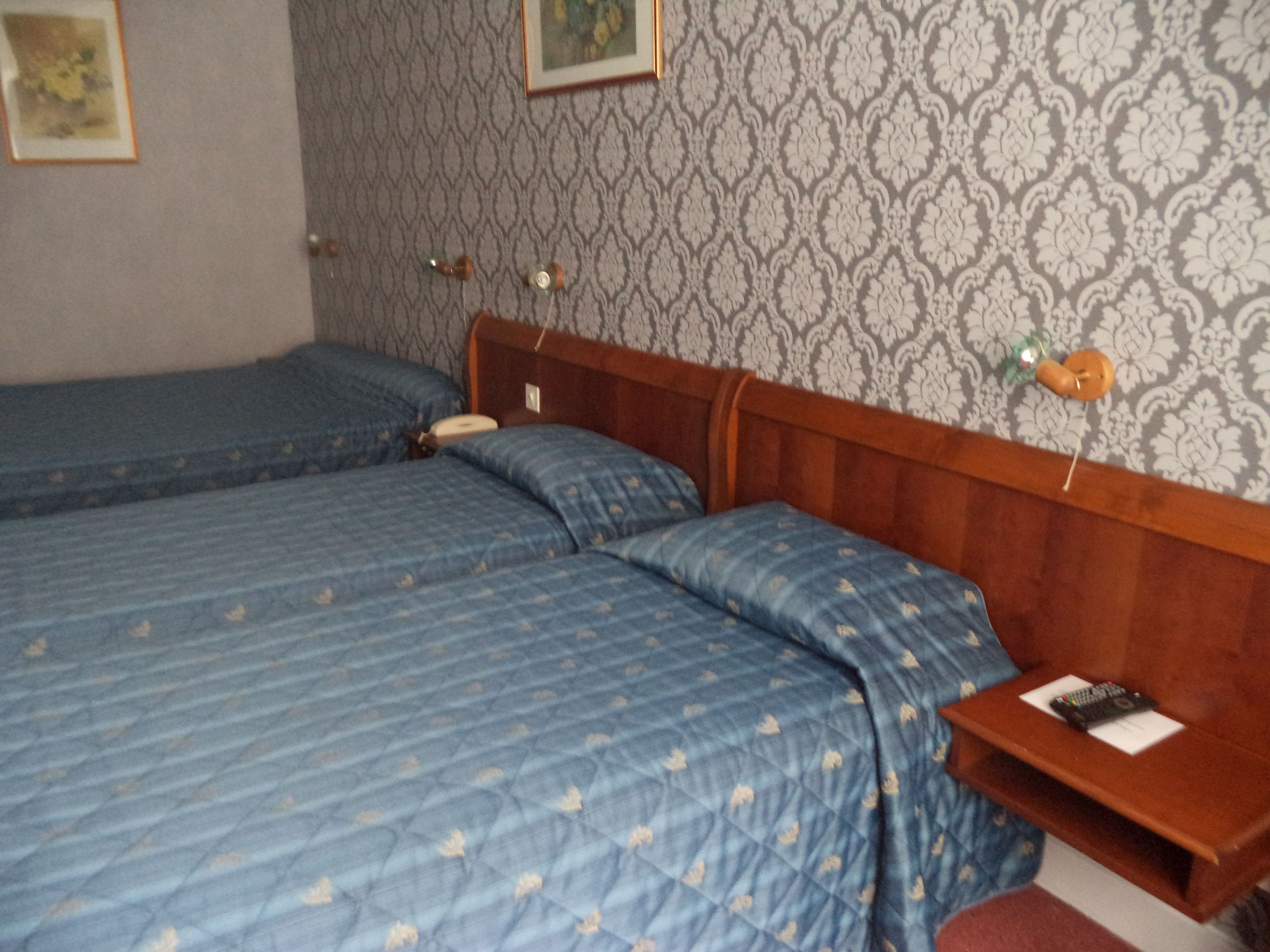 Habitación con dos camas, cabecero de madera, mesita de noche con lámpara y papel tapiz estampado.