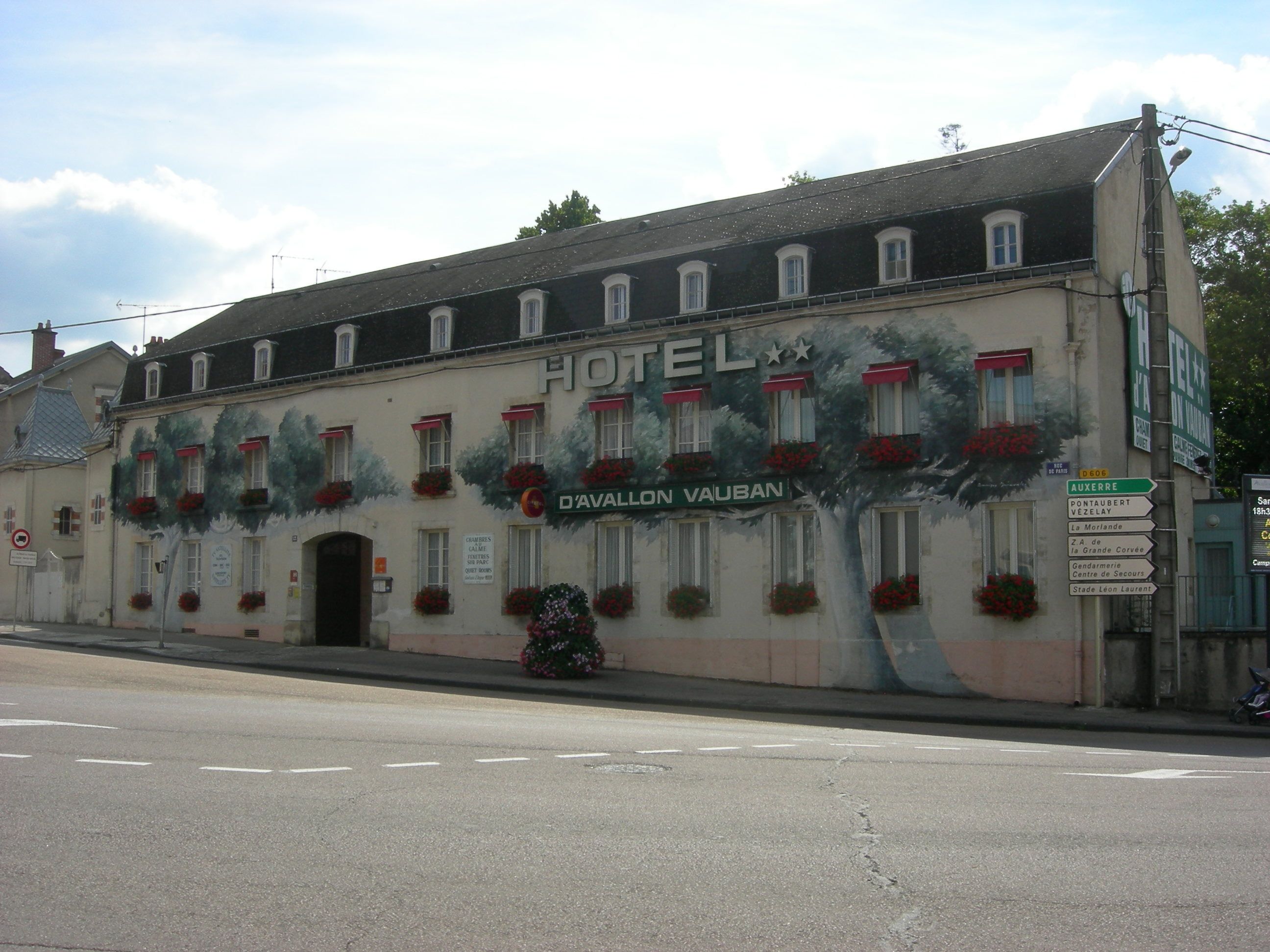 Een hotel met een muurschildering van bomen en bloemen op de gevel, een bordje met 'D'Avallon Vauban' erop, en een straatnaambordje met richtingaanduidingen.