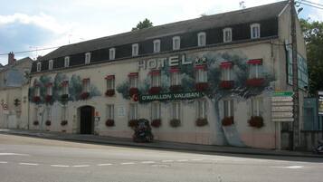 Een hotel met een muurschildering van bomen en bloemen op de gevel, een bordje met 'D'Avallon Vauban' erop, en een straatnaambordje met richtingaanduidingen.