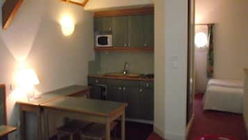 A compact hotel room with a kitchenette, a small table, a chair, a bed, and a nightstand.