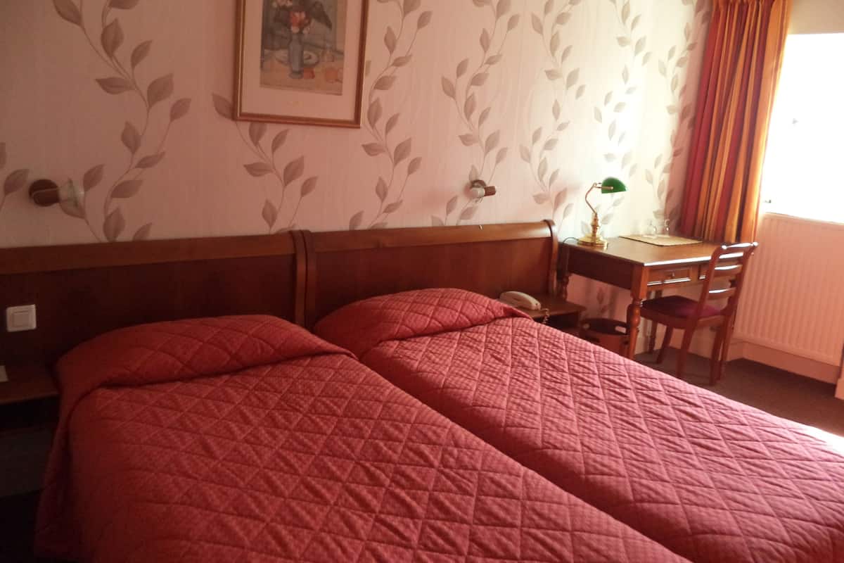 A hotel room with two beds, a desk, a chair, a lamp, and a window with curtains.