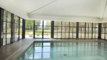 Una piscina interior con paredes de vidrio transparente, suelo de baldosas y vistas a un espacio exterior con un banco y árboles.