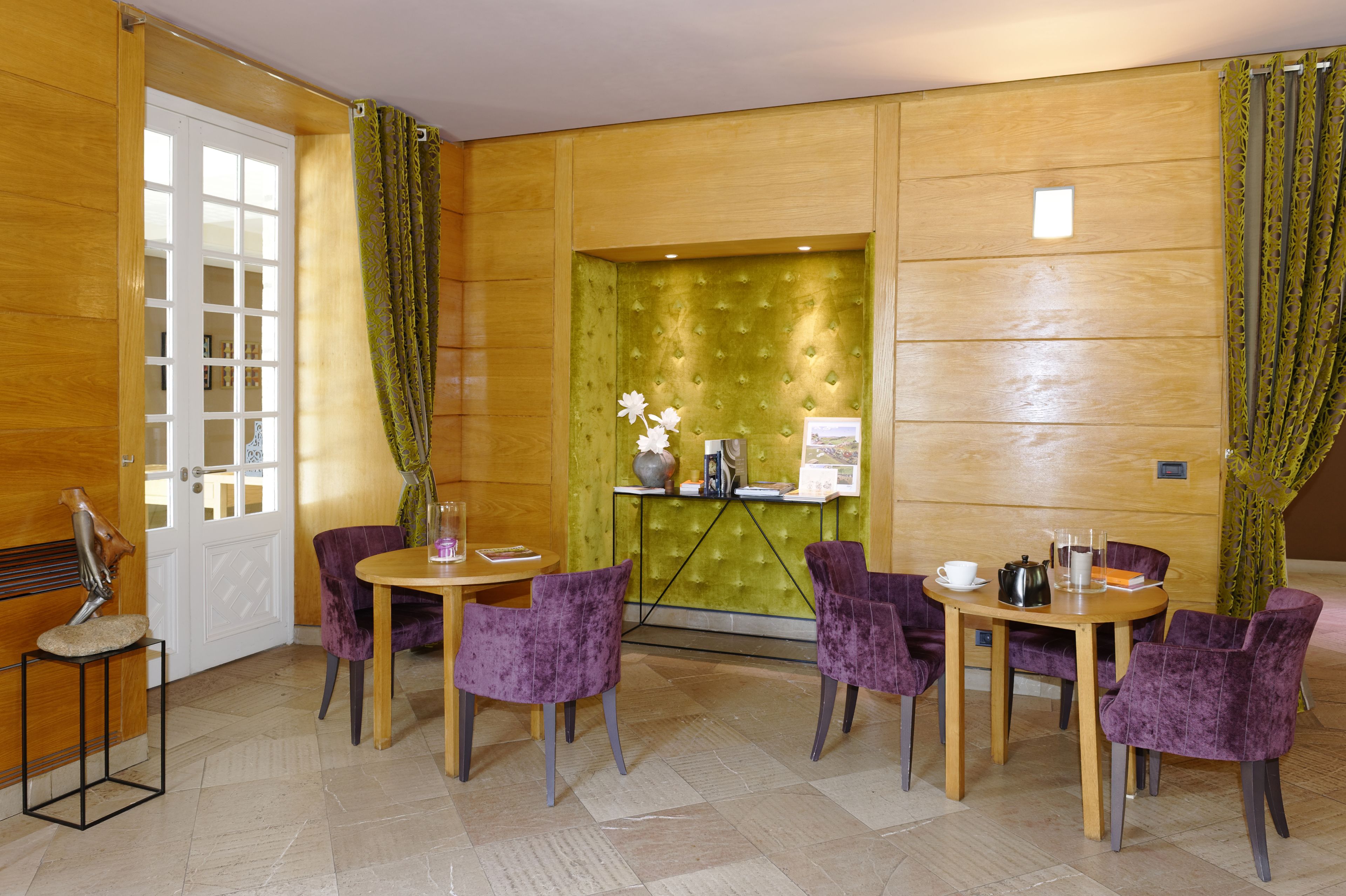 A hotel room with a wooden door, a green upholstered wall, a small table with a vase, and purple chairs around it.