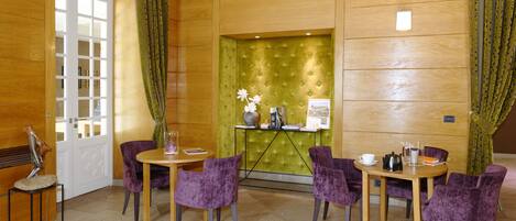 A hotel room with a wooden door, a green upholstered wall, a small table with a vase, and purple chairs around it.