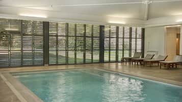 An indoor swimming pool with a tiled floor, a poolside area with lounge chairs, and a large window with blinds.