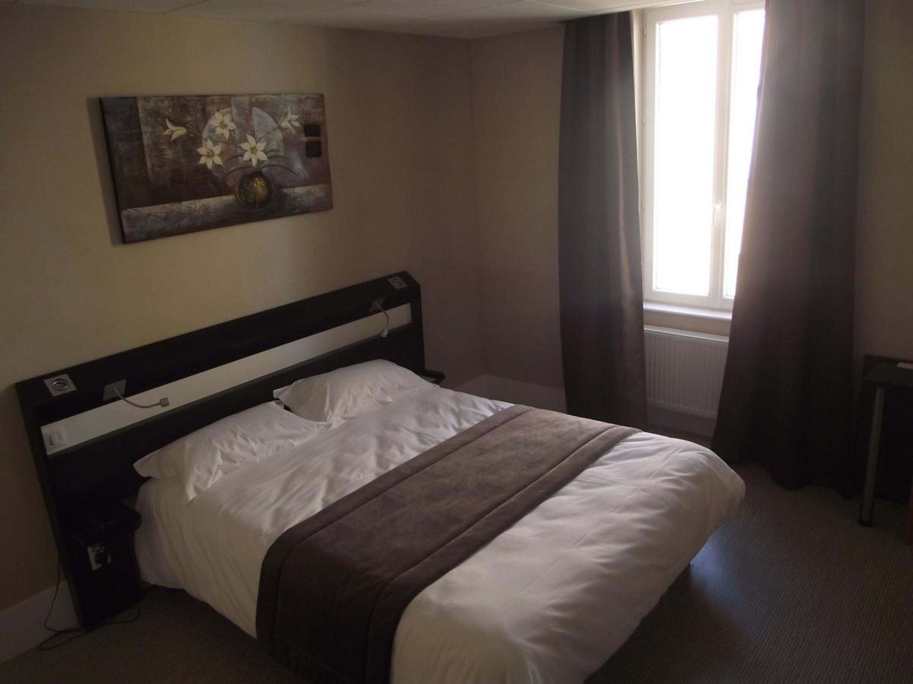 Una cama bien hecha en una habitación de hotel, con un cuadro en la pared, una ventana con cortinas y un radiador.