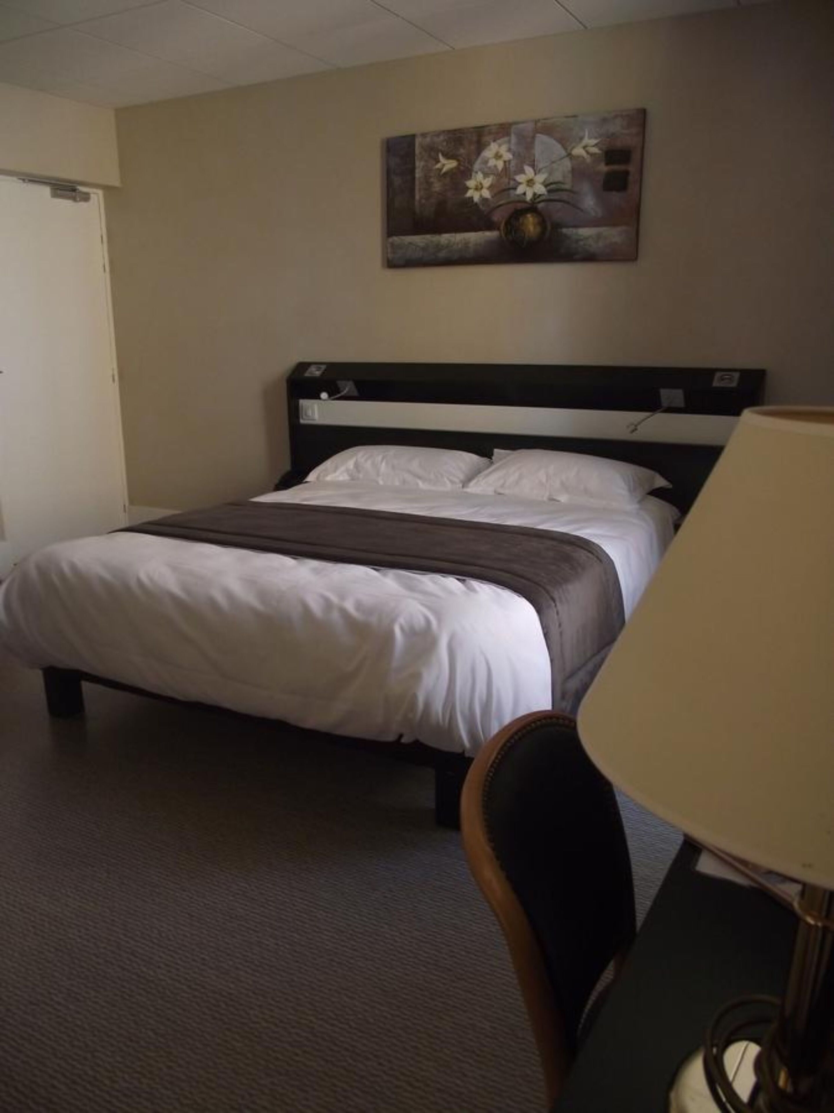 A hotel room with a bed, a chair, a desk, and a painting on the wall.