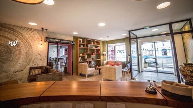 A modern hotel lobby with a wooden reception desk, comfortable seating, a bookshelf, and a view of the street through large glass doors. - Hôtel des Bourbons (Montlucon)