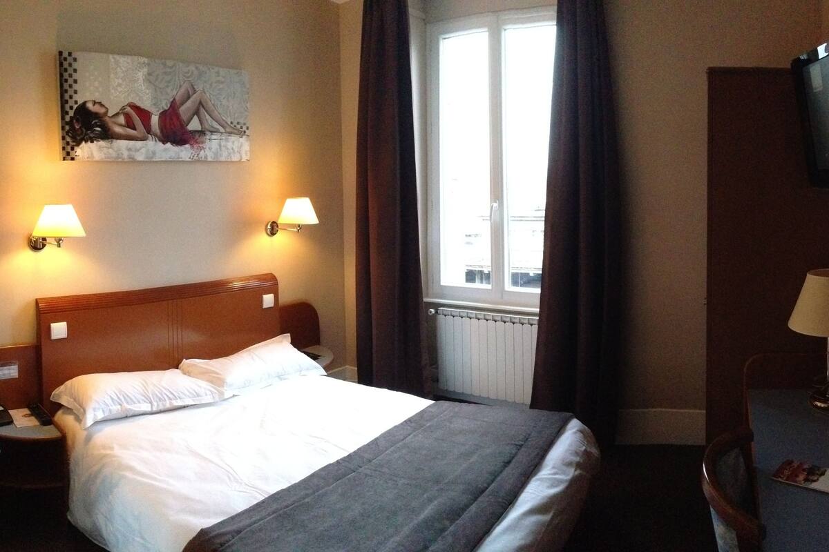 A hotel room with a bed, two bedside lamps, a chair, a desk with a lamp, a TV, and a window with curtains.