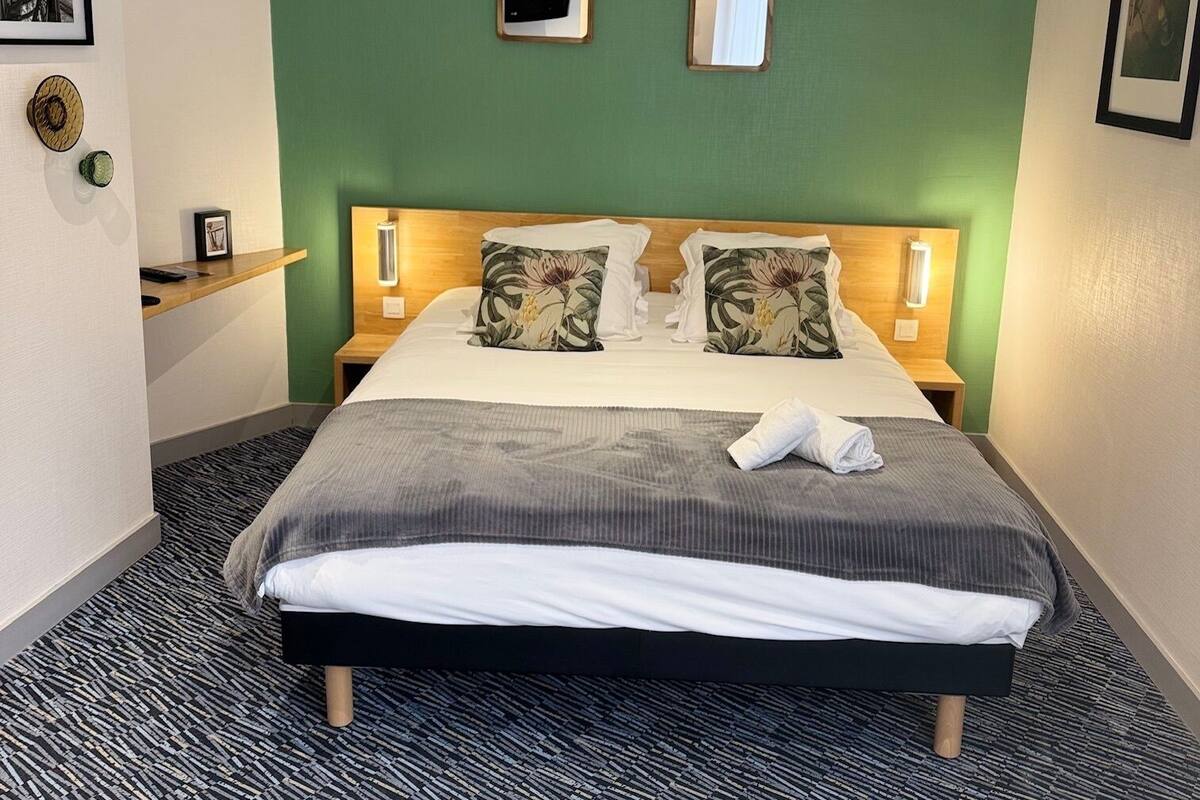 Quarto de hotel com uma cama grande, duas mesas de cabeceira, uma parede verde e uma pequena janela.