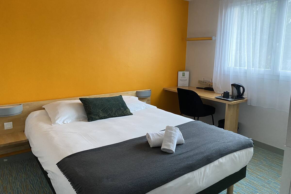 Quarto de hotel com cama, escrivaninha, cadeira, cafeteira e janela com cortinas.