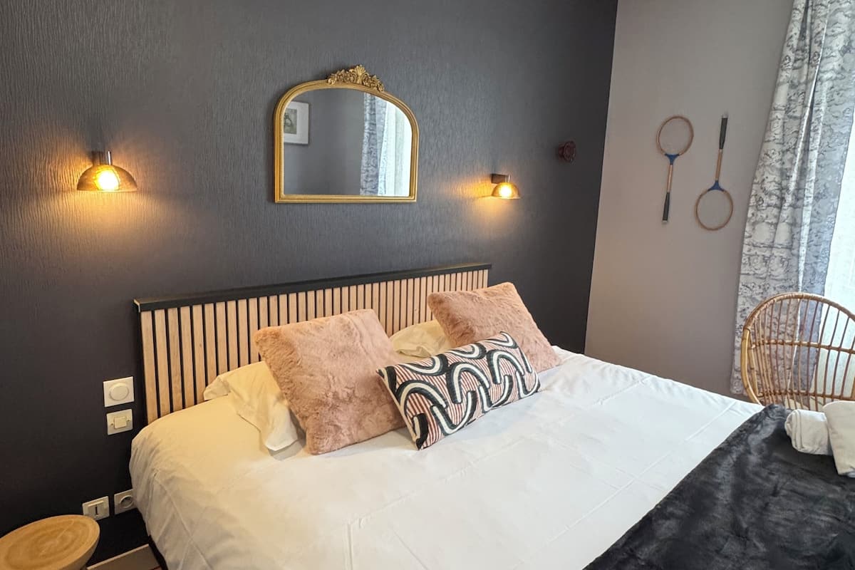 Uma cama bem arrumada com almofadas brancas e estampadas, um cobertor de cor preta e uma cabeceira com ripas verticais. Há duas luzes fixadas na parede e um espelho sobre a cama. Uma cadeira de vime é visível no canto, e o chão de madeira envolve o quarto.