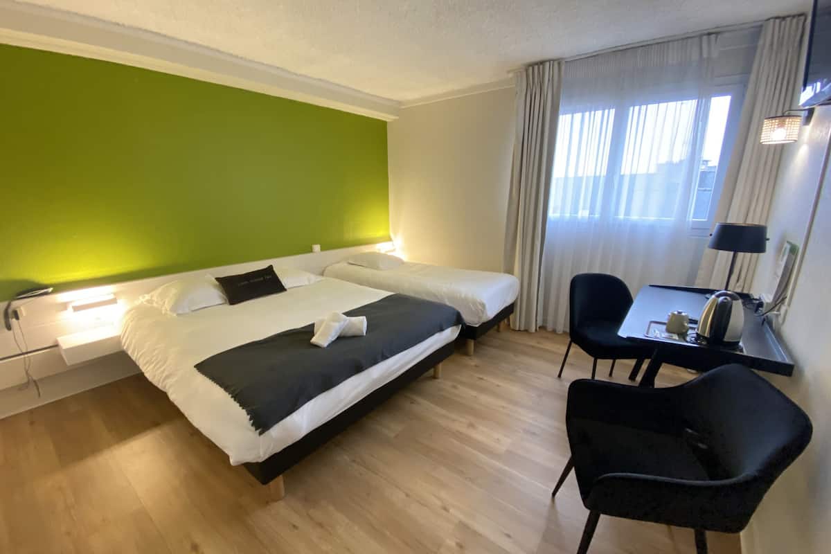 Quarto de hotel com duas camas, uma mesa de trabalho com cadeira, luminária e janela com cortinas.