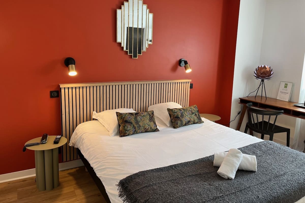 Quarto de hotel com cama, mesas de cabeceira, escrivaninha e luminária na parede.