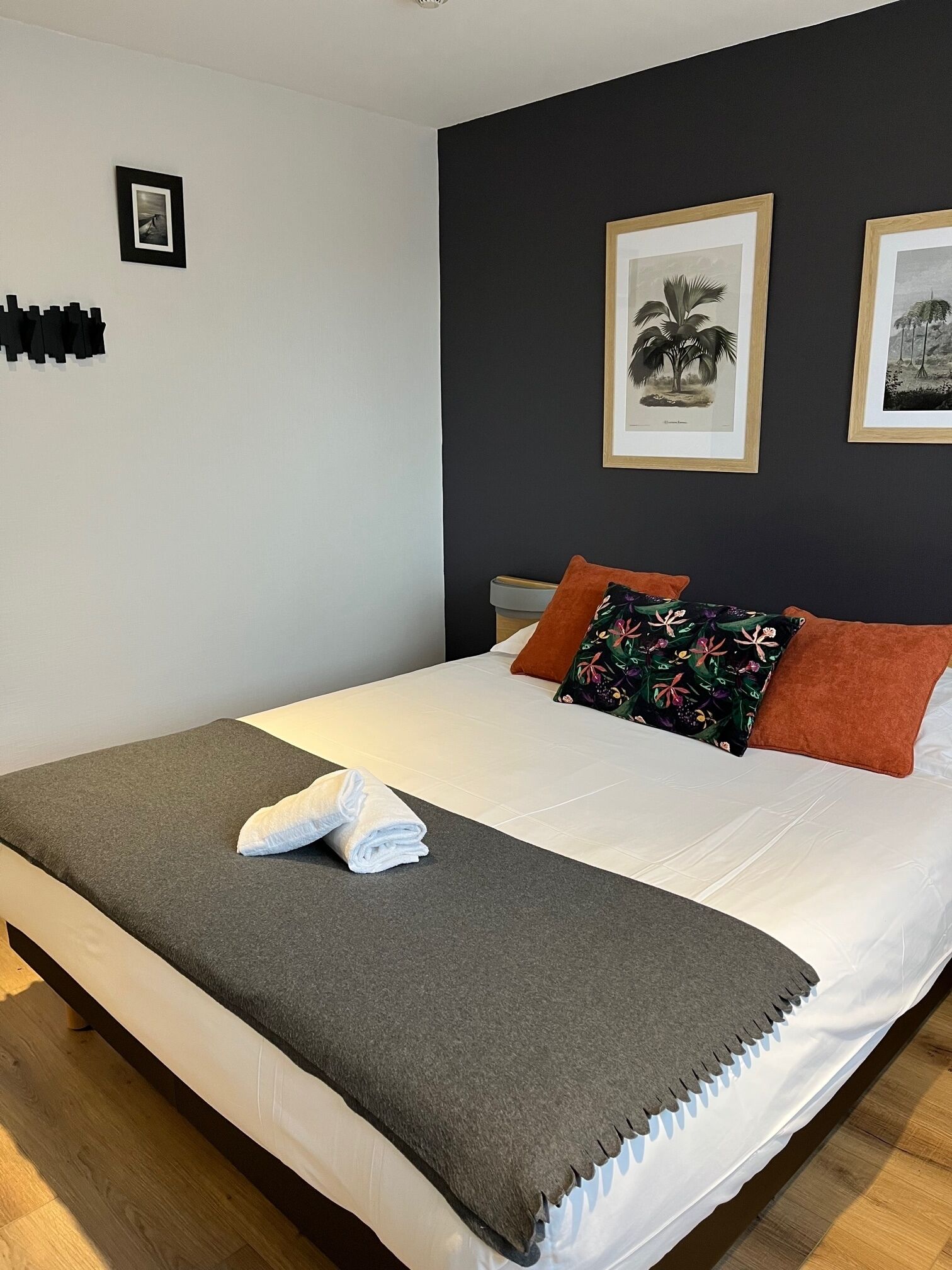 Ein ordentlich bezogenes Bett mit grauer Decke und einem gefalteten weißen Handtuch, zwei gerahmte botanische Drucke an der Wand und ein kleines schwarzes Kunstwerk links.