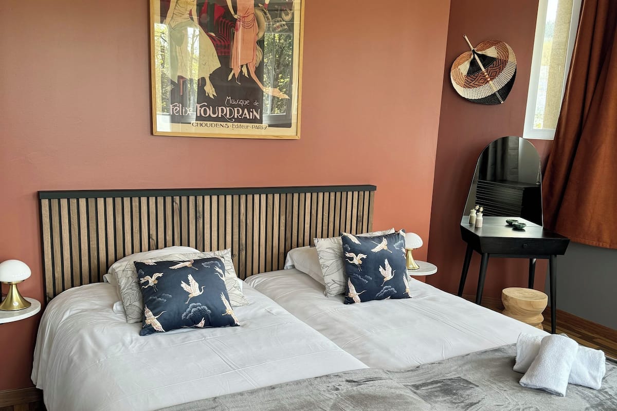 Uma cama de casal com duas almofadas, uma mesa de cabeceira com abajur, um espelho e um cartaz na parede.