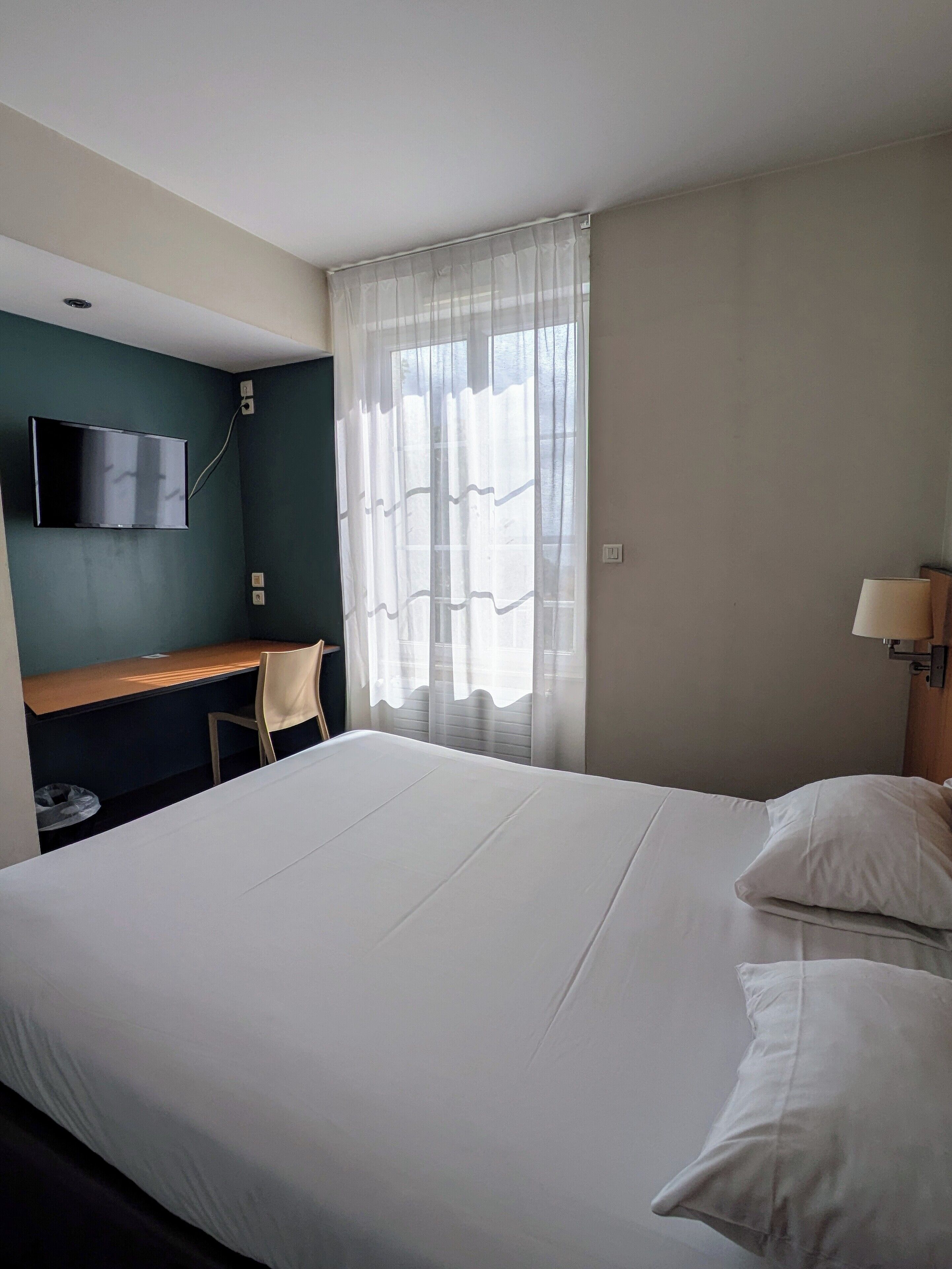 A hotel room with a bed, a desk, a chair, a TV, and a window with sheer curtains.