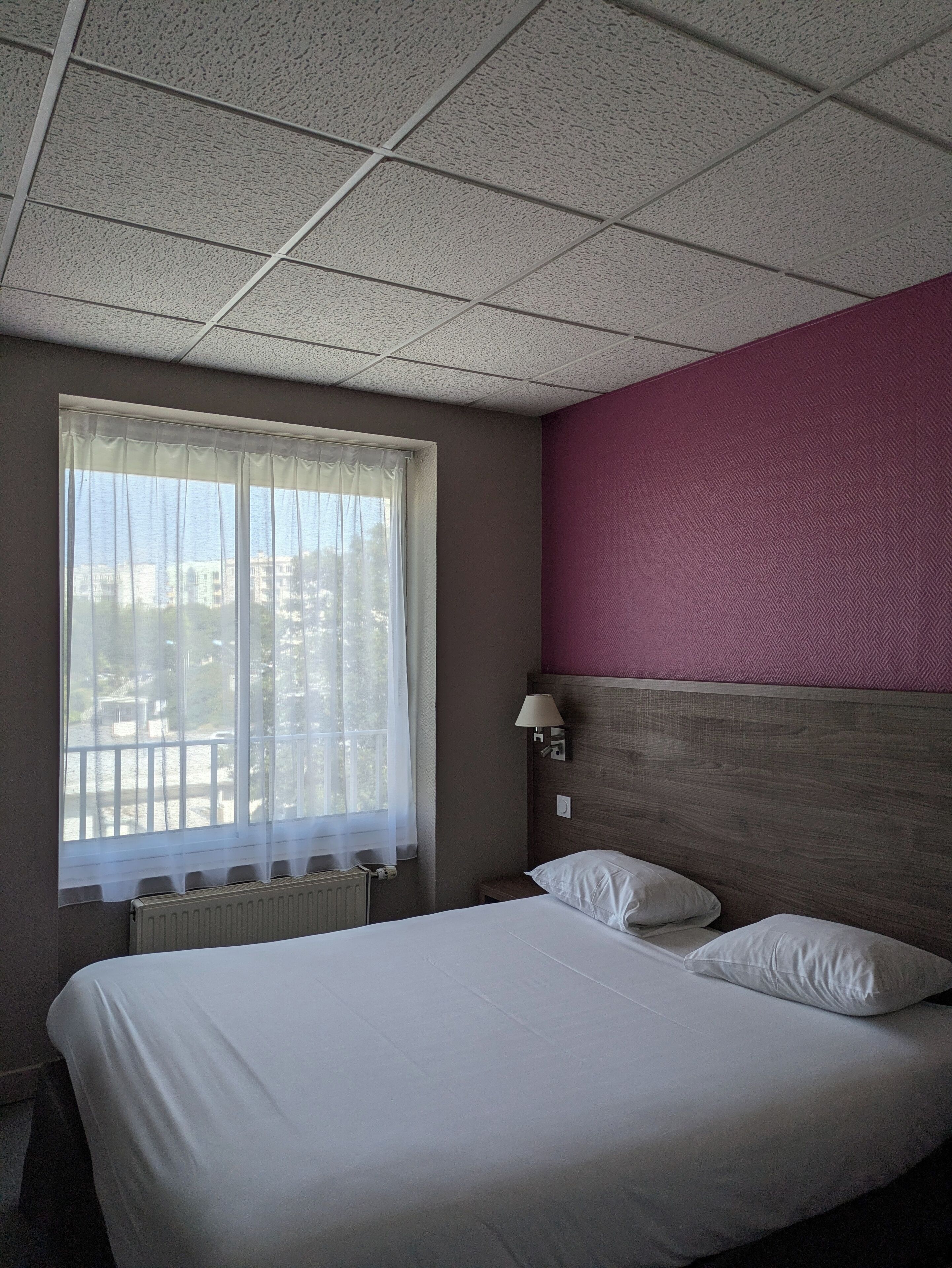A hotel room with a large bed, a window with sheer curtains, a wall-mounted lamp, and a radiator.