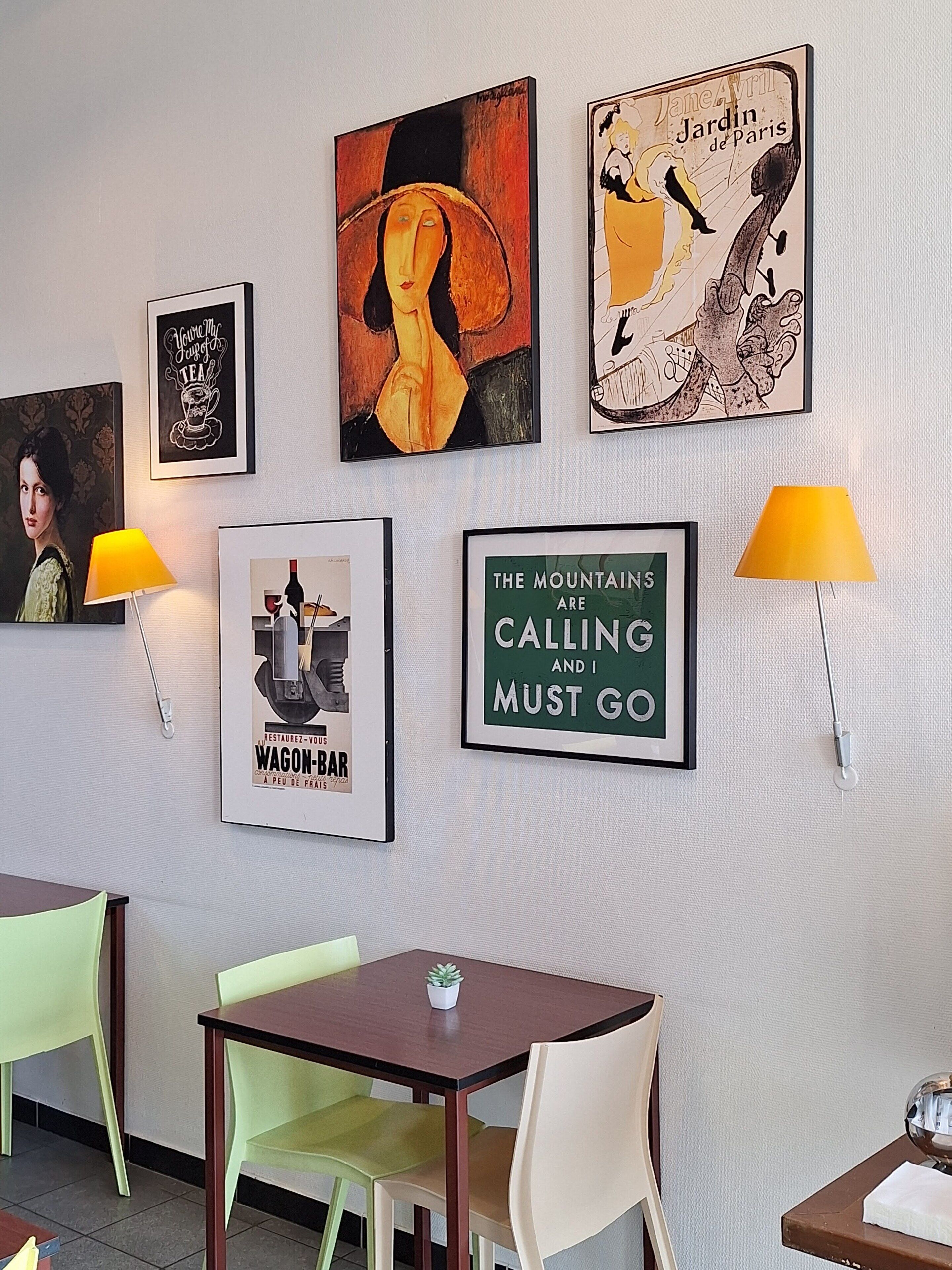 A wall with various framed artworks, a table with chairs, and two wall-mounted lamps.