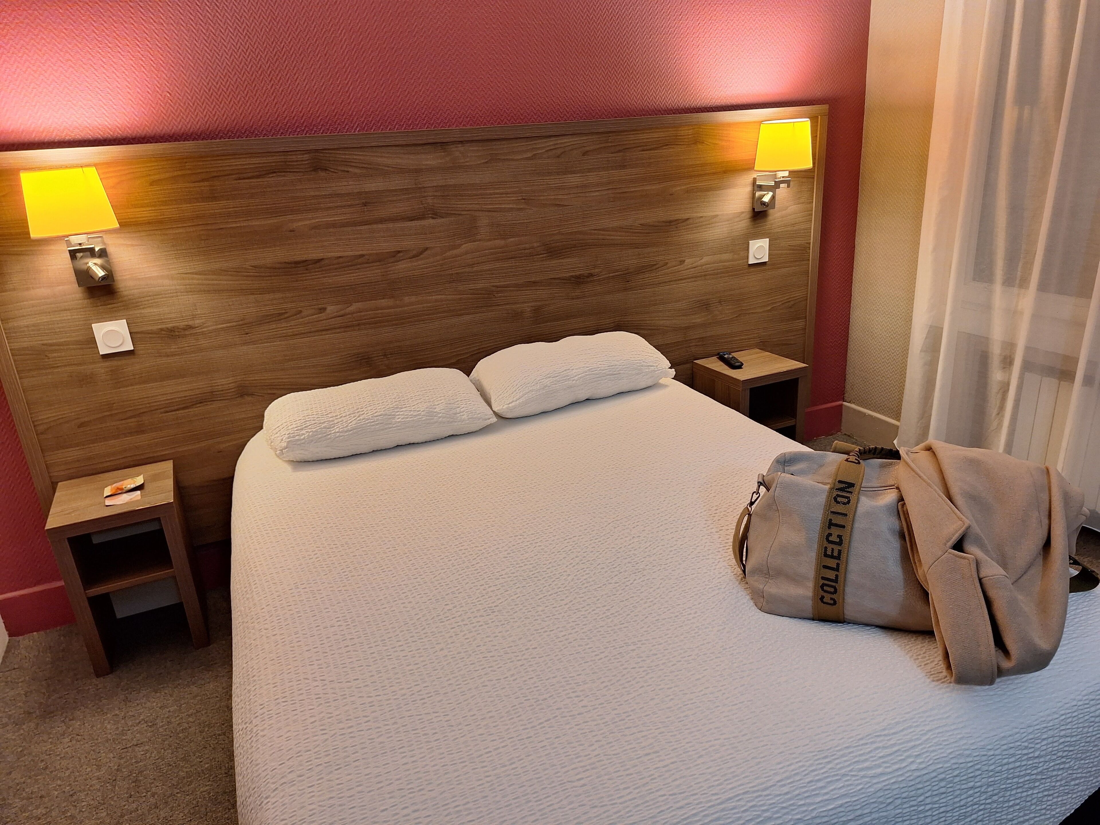 Een hotelkamer met een houten hoofdbord, twee bedlampen, een bed met witte beddengoed, een nachtkastje, een afstandsbediening en een tas op het bed.