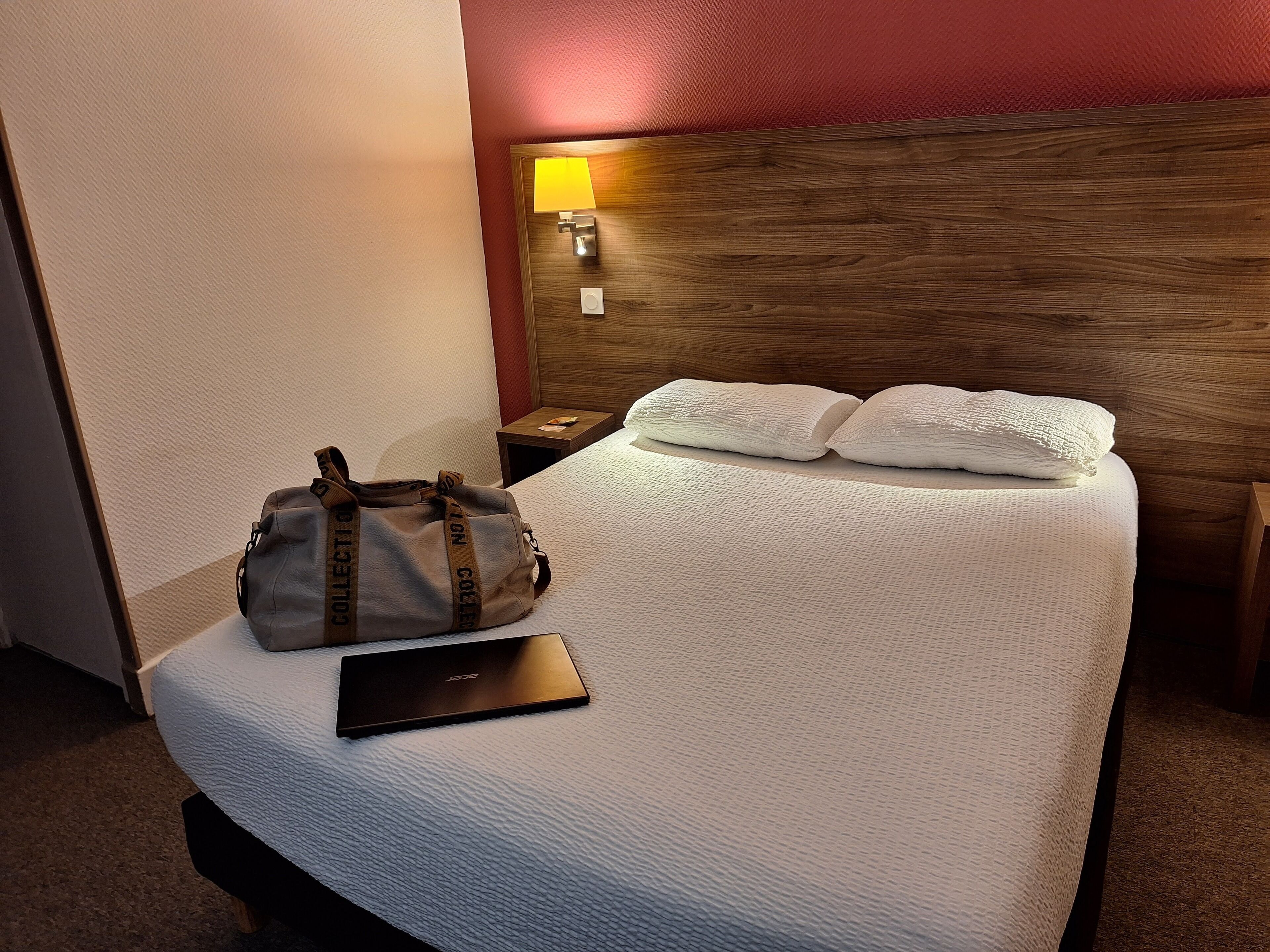 Un lit bien fait, avec des draps blancs, une tête de lit en bois, une lampe de chevet, un ordinateur portable noir et un sac marron posé sur le lit.