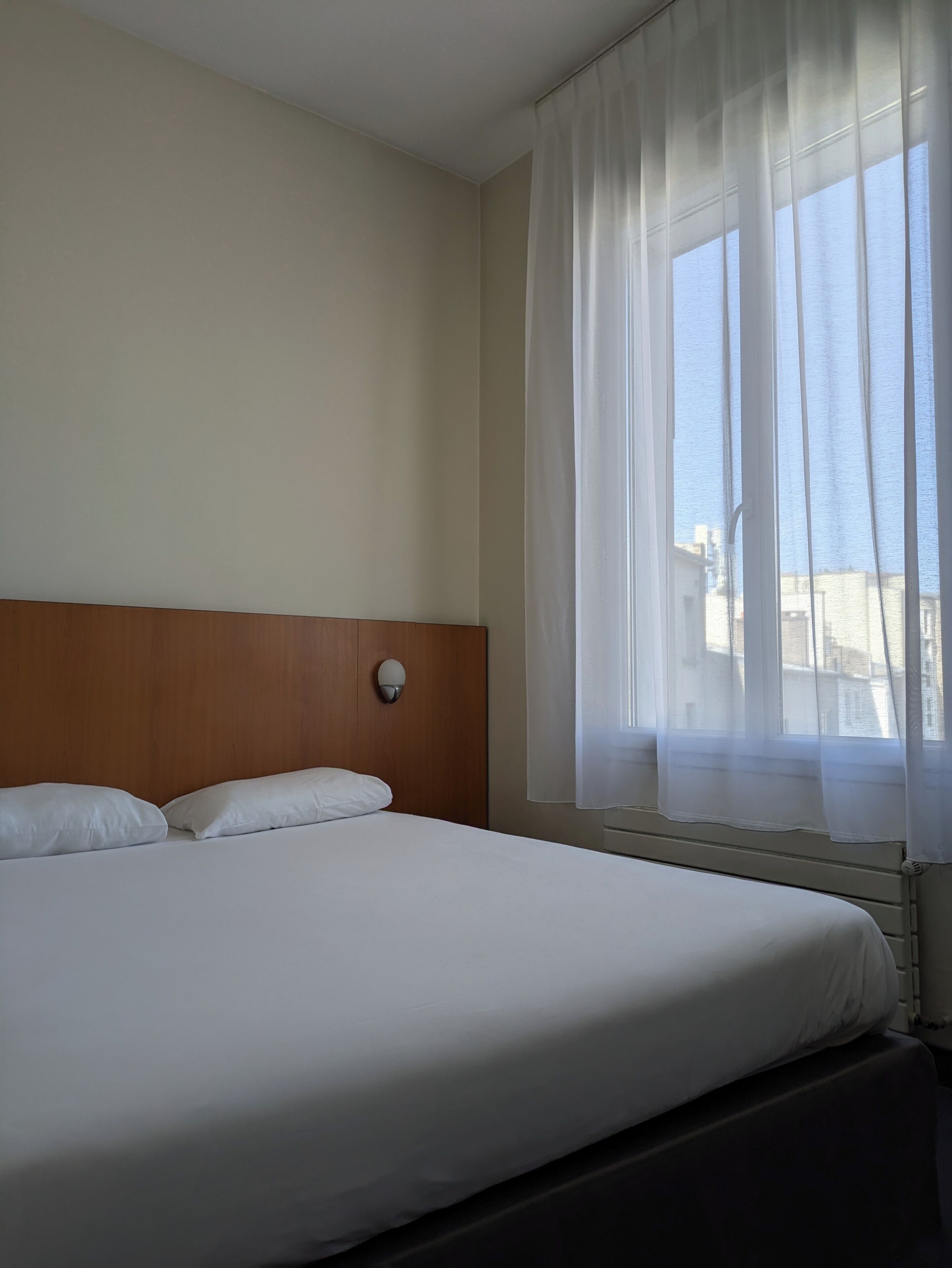 A hotel room with a large bed, a wooden headboard, a window with sheer curtains, and a view of buildings outside.